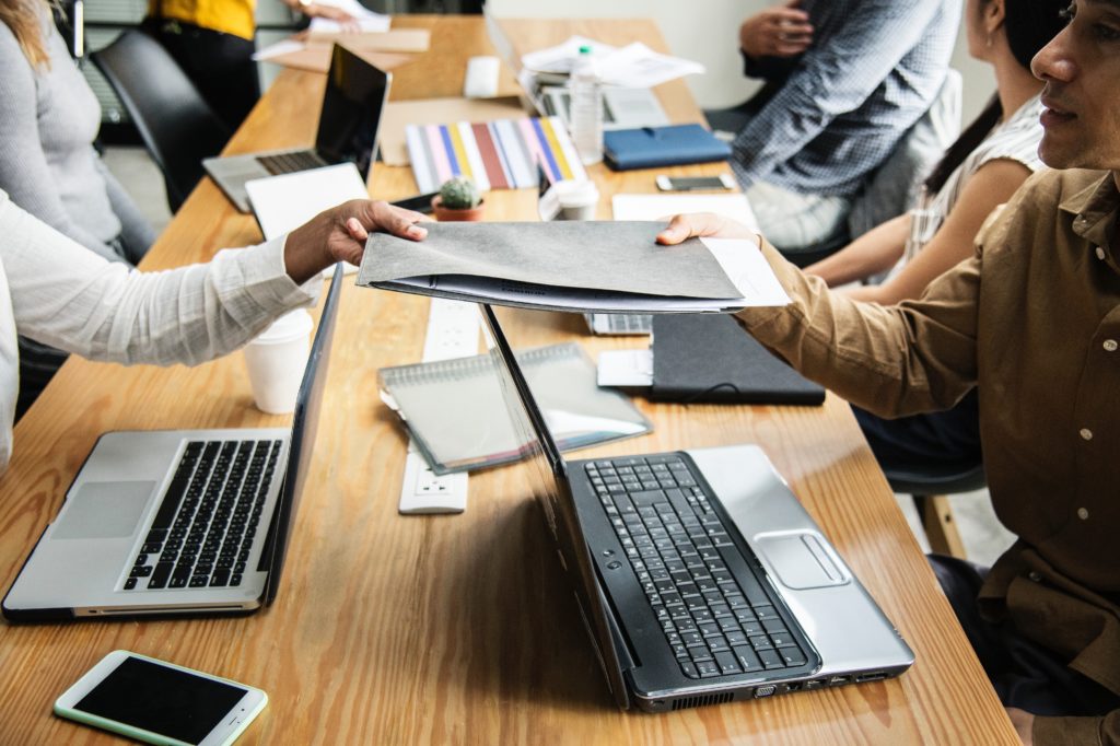 one person hands a file to another over a laptop
